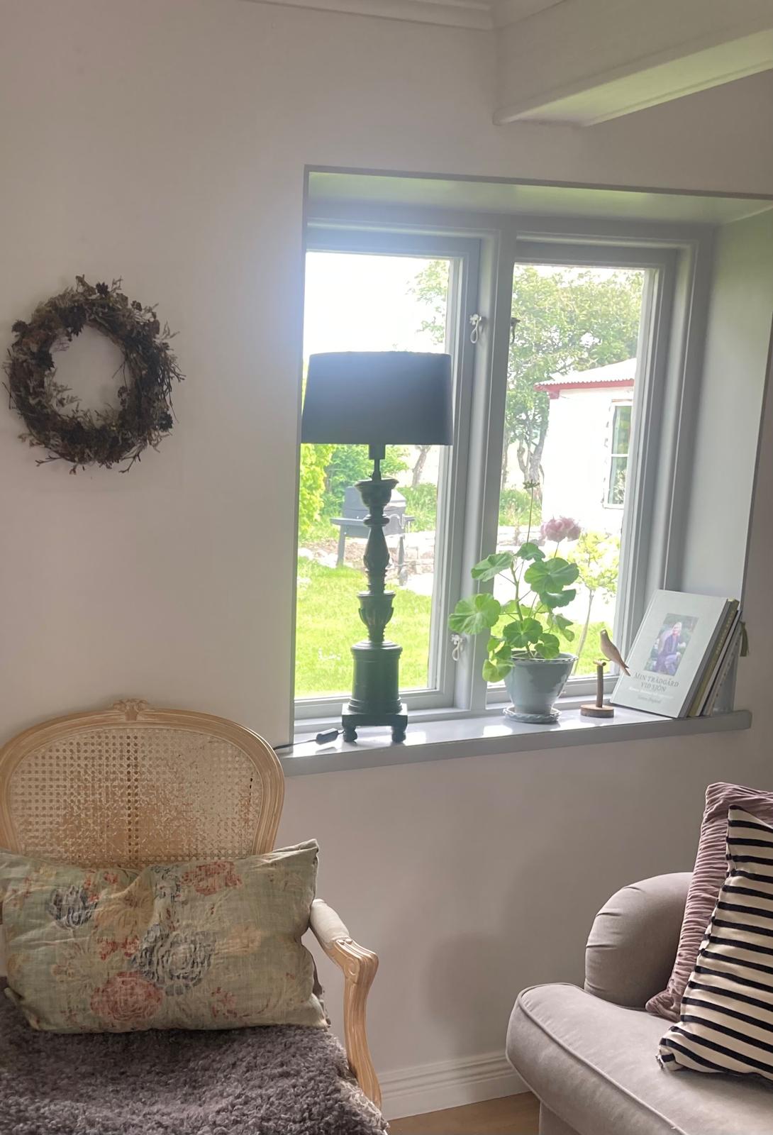 Rottingfåtölj, torkad krans på väggen och fönsterbräda med lampa och växter